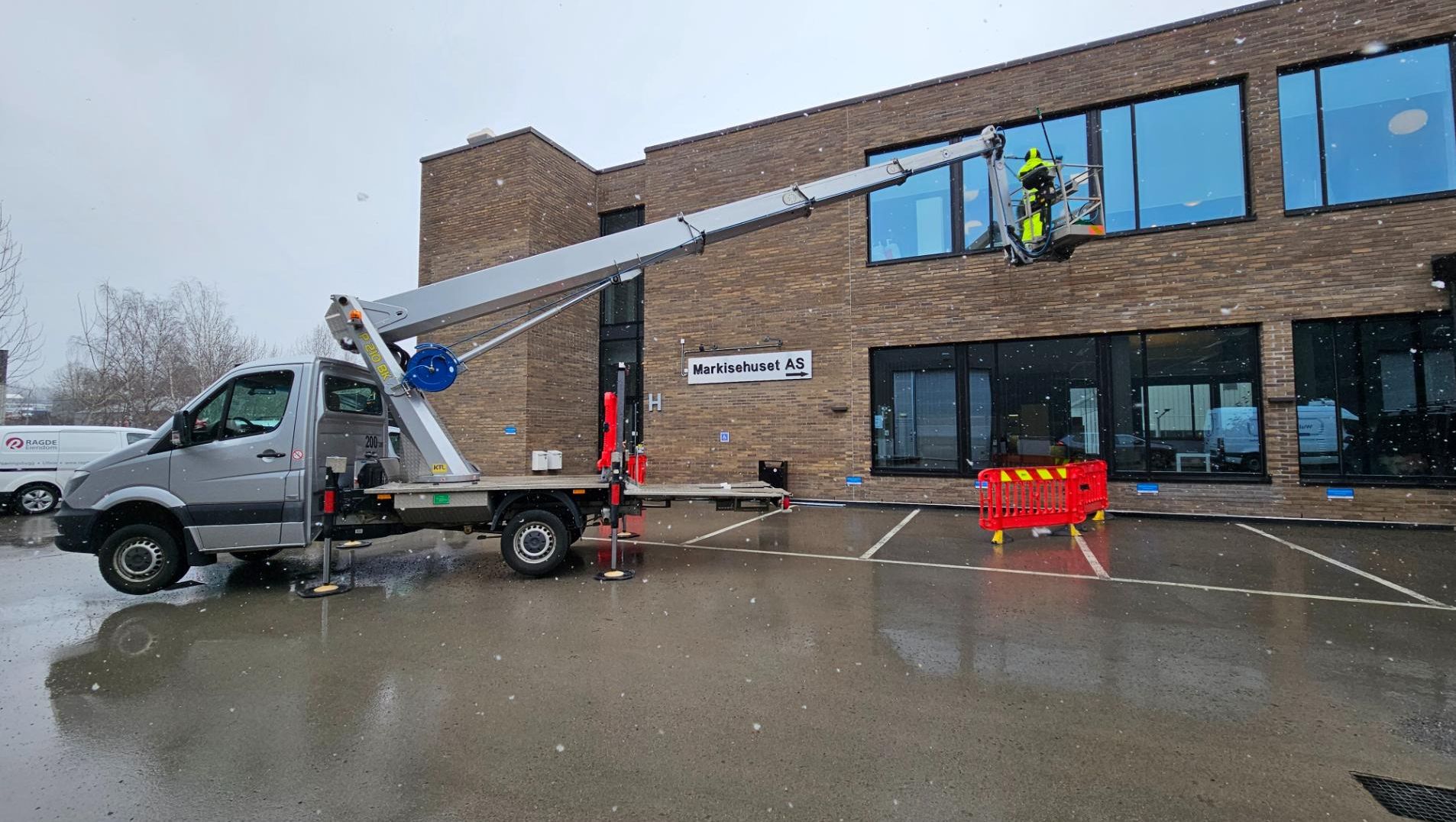 Grå liftbil med støtteben og kurv i arbeid foran murbygning i regnvær med snøfnugg i luften.