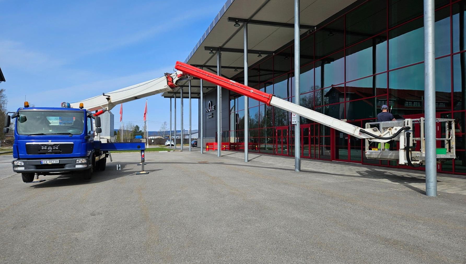 Liftbil med hevet arm og arbeider i kurv foran bygning med store glassvinduer og røde karmer.
